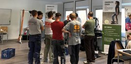 Vorführung Exoskelett Eine Gruppe junger Männer stehen im Kreis und bekommen ein Exoskelett erklärt