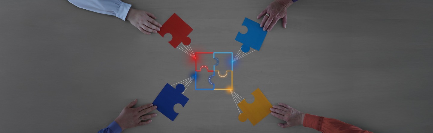 Zusammenarbeit für eine vollständige Lösung mit Puzzleteilen Vier Hände die jeweils Puzzleteile halten. Diese werden in der Mitte des Bildes zusammengefügt.