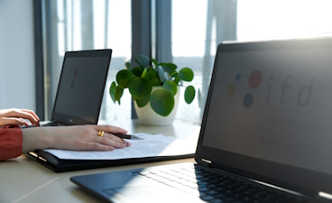 Zwei Laptops stehen auf einem Tisch und jemand tippt auf dem linken Laptop. Eine Akte liegt daneben.