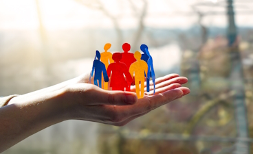 2 Hände sind nach oben offen ausgestreckt. Darauf ist ein Kreis mit menschlichen, bunten Umrissen.