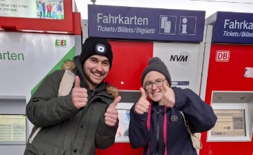 Zwei gut gelaunte, junge Menschen kaufen Fahrkarten am Bahnhof.